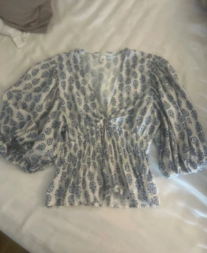 Vit blus med blått mönster och puffärm - Söt vit blus med blått mönster och puffiga, trekvartsärmar. Djup v-ringning och smockad midja.    Fint skick, storlek XS