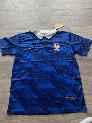 Blå Frankrike Nike fotbollströja - Snygg blå fotbollströja från Nike med Frankrikes landslagsemblem och vita detaljer. Tröjan har korta ärmar, vit krage och ett diagonalrandigt mönster i olika blå nyanser. Perfekt för match eller träning.