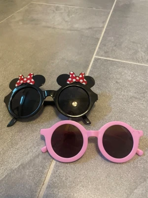 Två par runda solglasögon för barn - Säljer två par solglasögon med rund form för barn. Ett par är svart med öron och röda prickiga rosetter, inspirerade av Musse Pigg. Det andra paret är helrosa med enkel design. Båda är perfekta för dig som vill sticka ut i sommar.