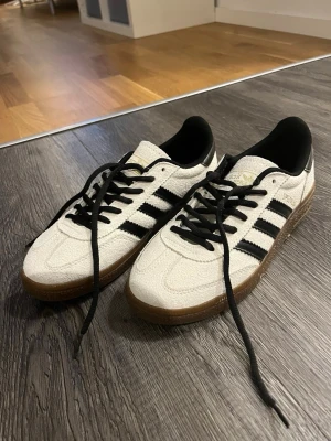 Adidas Spezial vita sneakers - Säljer ett par Adidas Spezial sneakers i vitt med svarta ränder och detaljer. Skorna har klassisk låg profil, svart innersula och gummisula. Materialet är mocka och textil, med svarta skosnören och gulddetaljer på sidan och plösen.(har använts 1 gång köpets för 2 veckor sen)