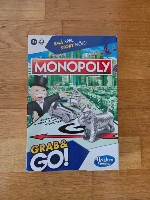 Monopoly Grab & Go sällskapsspel - Monopoly Grab & Go är ett kompakt sällskapsspel med klassiskt Monopol-tema. Spelet har en färgglad kartong i blått, grönt och vitt och innehåller spelplan, spelpjäser i metall, kort och pengar. Perfekt för att ta med på resan eller spela hemma med vänner. För 2-4 spelare, från 8 år.
