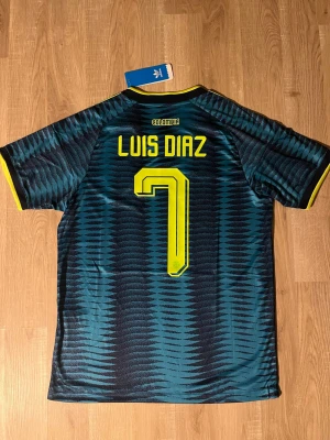 Colombia VM-fotbollströja Luis Diaz #7 - Snygg Colombia fotbollströja med Luis Diaz och nummer 7 på ryggen. Tröjan är mörkblå med gula detaljer, mönstrad design och klassiska ränder på axlarna. Tillverkad i lätt och ventilerande material, perfekt för träning eller match. HELT NY!