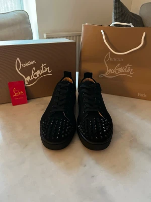 Svarta sneakers med nitar från Louboutin - Säljer ett par svarta sneakers från Christian Louboutin med coola svarta nitar på tån och den ikoniska röda sulan. Skorna har snörning och är tillverkade i skinn för en lyxig känsla. Perfekta för dig som vill sticka ut med din stil.