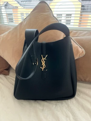 Svart bucket bag från Saint Laurent - Stilren svart bucket bag i läder från Saint Laurent med justerbar axelrem och ikoniskt YSL-spänne i guld framtill. Väskan har en mjuk, strukturerad form och elegant design som passar perfekt till din outfit.