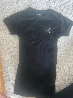 Svart t-shirt från Peach - En svart t-shirt från Peach med rund hals och korta ärmar. Diskret logga med texten 'peach' och en liten persika på bröstet. Tillverkad i mjuk bomull som känns skön mot huden. Perfekt till jeans eller shorts.