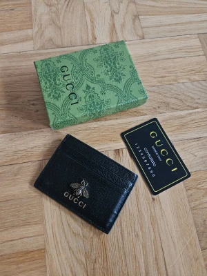 Svart korthållare med Bi - Snygg svart korthållare i läder med guldfärgad bi-detalj och logotyp framtill. Kommer med original grön ask och äkthetskort. Perfekt för att hålla koll på dina kort med stil.