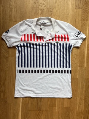 Vit pikétröja från Lacoste med ränder - Helt ny, Snygg vit pikétröja från Lacoste med röda och marinblå vertikala ränder på bröstet och svarta ränder nedtill. Klassisk krage och kort ärm med tryck på båda ärmarna. Ikoniska krokodilloggan på bröstet. Tillverkad i pikématerial.