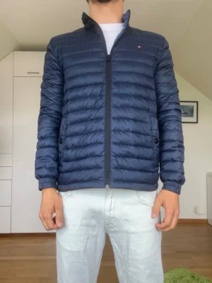 Mörkblå dunjacka från Tommy Hilfiger - Mörkblå dunjacka från Tommy Hilfiger. Det följer med en påse där jackan får plats i (se bild 6) strl S. Inga defekter. Modellen är 178 cm och väger 77 kg. Hör gärna av dig vid frågor!