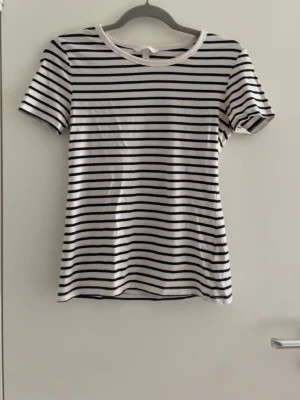 Randig t-shirt med marinblå ränder - Säljer en klassisk vit t-shirt med tunna marinblå horisontella ränder. T-shirten har rund halsringning och korta ärmar. Perfekt för en enkel och stilren look. Materialet ser ut att vara mjukt och bekvämt, troligtvis bomull.