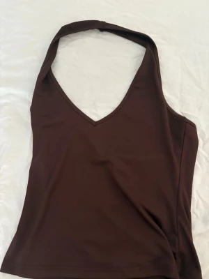 Brun halterneck linnetopp - Snygg brun linnetopp med halterneck och v-ringad urringning. Toppen är ärmlös och har en tight passform som framhäver axlarna. Perfekt att styla med jeans eller kjol för en trendig look.