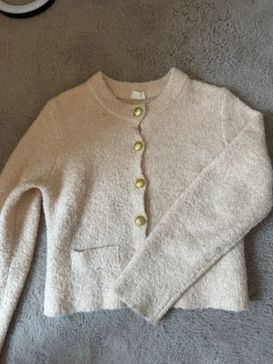 Beige kofta med guldfärgade knappar - Mysig beige kofta med rund halsringning och fyra dekorativa guldfärgade knappar framtill. Koftan har en liten ficka på ena sidan och är tillverkad i ett mjukt, strukturerat material. Perfekt för lager på lager och enkel att matcha.