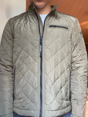 Quiltad beige jacka från Replay - Snygg quiltad jacka från Replay i beige med dragkedja framtill och ståkrage. Jackan har diskreta fickor med dragkedja, bröstficka och Replay-logga på ärmen. Perfekt för höst och vår, med en modern och stilren look.