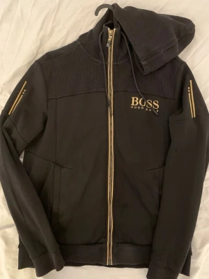 Svart hoodie från Hugo Boss - Snygg svart hoodie från Hugo Boss med dragkedja och guldfärgade detaljer på ärmar och dragkedja. Stort BOSS-tryck på bröstet och huva med dragsko. Insidan är beige och materialet känns mjukt och bekvämt. Perfekt för en chill och stilren look.
