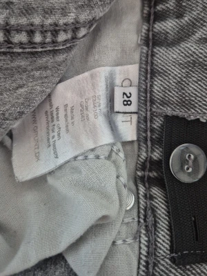 Grå jeans med råa kanter, storlek 28 - Säljer ett par grå jeans i storlek 28 med trendiga råa kanter nertill. Jeansen har en klassisk femficksmodell och stängs med knapp och dragkedja. Materialet är mjukt och följsamt, perfekt för en avslappnad look. Lite slitna i nederkant (se bild)