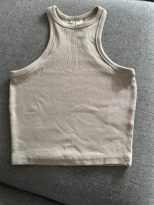 Beige ribbad croppad linnetopp - Säljer en beige ribbad croppad linnetopp med hög hals och ärmlös design. Perfekt till sommaren och enkel att matcha med olika stilar. Mjuk och stretchig känsla i materialet.