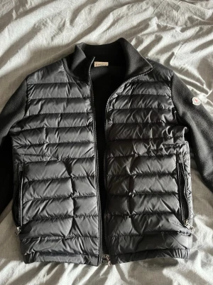 Moncler cardigan  - Snygg svart dunväst från Moncler med quiltad framsida och två dragkedjefickor. Västen har stickade ärmar och rygg, samt hög krage för extra värme. Klassisk Moncler-logga på ärmen och dragkedja framtill. Perfekt för lager-på-lager och streetstyle.