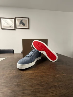 Christan Louboutin  - Snygga blå sneakers i mocka med vit mellansula och ikonisk röd undersida. Skorna har klassisk låg profil och stilrena snörningar. Perfekta för dig som vill sticka ut med en unik detalj under foten.