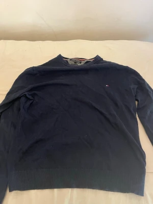 Mörkblå stickad tröja Tommy Hilfiger - Mörkblå stickad tröja från Tommy Hilfiger med rund halsringning och diskret logga på bröstet. Tröjan har långa ärmar och ribbade muddar vid ärmslut och nederkant. Perfekt till höst och vinter för en stilren look.