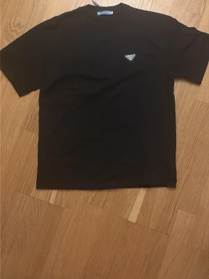 Svart t-shirt från Prada - Snygg svart t-shirt från Prada med diskret logga på bröstet. Klassisk rund hals och korta ärmar. Tillverkad i mjuk bomull som känns skön mot huden. Perfekt för dig som gillar stilrena och exklusiva plagg. Står storlek S men passar mer som en M