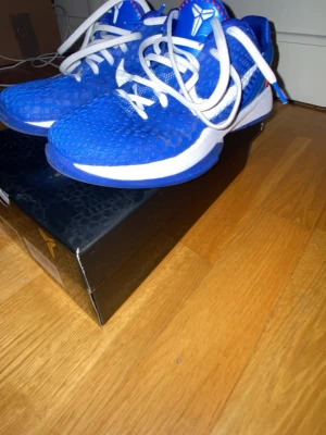 Blå Nike Kobe sneakers - Säljer ett par blå Nike Kobe sneakers med vit sula och vita detaljer. Ovandelen har ett mönstrat, nästan reptil-liknande material och skorna har vita skosnören. Kobe-loggan syns tydligt på plösen och sulan. Perfekta för dig som gillar basket och streetwear.