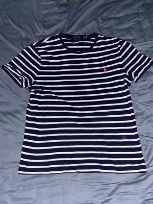 Randig t-shirt från Polo Ralph Lauren - Snygg marinblå och vit randig t-shirt från Polo Ralph Lauren med klassisk passform. T-shirten har korta ärmar och en liten röd logga broderad på bröstet. Perfekt till jeans eller shorts för en clean look.