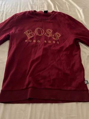  Hugo Boss hoodie - Snygg vinröd sweatshirt från Hugo Boss med stor gul logga på bröstet. Tröjan har rund halsringning, ribbade muddar vid ärmslut och nederkant samt en klassisk passform. Tillverkad i mjukt bomullsmaterial för skön känsla. Tröjan är i storlek XL men passar M