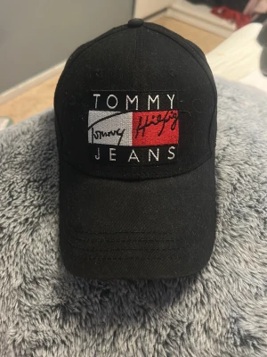 Svart keps från Tommy Jeans - Säljer en svart keps från Tommy Jeans med broderad logga framtill i vitt, rött och svart. Klassisk böjd skärm och justerbar rem bak. Materialet är bomull och kepsen har en stilren och sportig look.