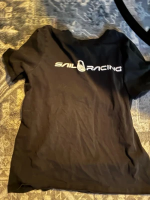 Svart Sail Racing t-shirt - Svart t-shirt från Sail Racing med vit logga tryckt över bröstet. Klassisk passform och rund halsringning. Perfekt för dig som gillar enkel och stilren design. Tillverkad i mjuk bomull för skön känsla hela dagen.