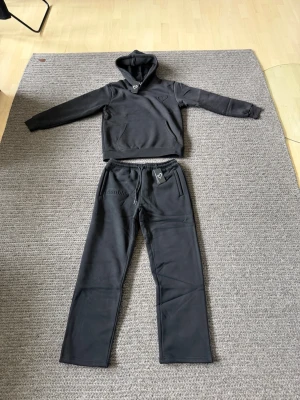 Svart loungewear-set från Doublés - Mysigt svart loungewear-set från Doublés med hoodie och matchande byxor. Hoodien har ficka framtill och byxorna har dragsko i midjan samt broderad logga. Perfekt för chill dagar hemma. Mjukt material som känns skönt mot huden.