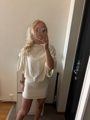 Beige kort klänning med vida ärmar - Säljer en beige kort klänning med lös passform upptill och vida, halvlånga ärmar. Kjoldelen är tightare och markerar midjan snyggt. Klänningen har en enkel, stilren design och är tillverkad i ett mjukt, lätt material som ger ett elegant intryck. Aldrid använd!
