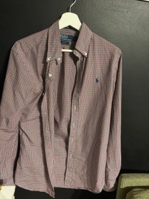 Rutig skjorta från Polo Ralph Lauren - Klassisk rutig skjorta från Polo Ralph Lauren i rött, blått och vitt. Skjortan har lång ärm, knappar framtill och button-down krage. Tillverkad i mjuk bomull och har den ikoniska loggan broderad på bröstet. Perfekt för en stilren och avslappnad look.