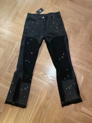 Svarta jeans med färgstänk och rå kant - Unika svarta jeans med vita färgstänk och råa, fransiga benslut. Jeansen har raka ben och patchade detaljer vid knäna. Tillverkade i denim med en cool, avslappnad vibe. Perfekta för dig som vill sticka ut med din stil.