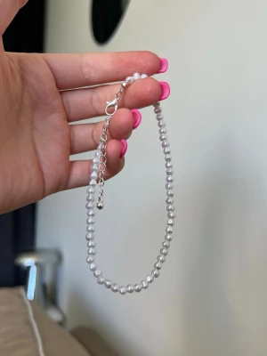 Fotlänk med små vita pärlor - Söt fotlänk med små vita pärlor och justerbart silverfärgat lås. Pärlorna är runda och stilrena. Kan passa både som ankelkedja eller armband beroende på hur du vill bära det.
