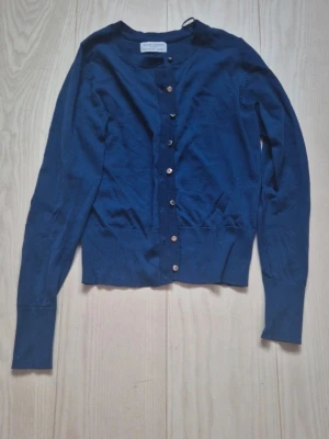 Mörkblå cardigan från Holly & Whyte - Mörkblå cardigan från Holly & Whyte med rund halsringning och guldiga knappar framtill. Långärmad modell med ribbade muddar och nederkant. Perfekt att slänga över en t-shirt eller klänning för en snygg lager-på-lager-look.