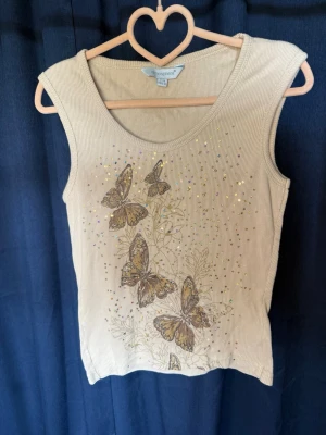 Beige ribbad linnetopp med fjärilar - Snygg beige ribbad linnetopp med glittriga paljettdetaljer och tryck av bruna fjärilar framtill. Perfekt för dig som vill ha en unik och somrig topp med lite extra bling. Ärmlös och med rund halsringning.