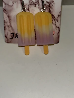 Glasspinne örhängen i pastellfärger - Säljer ett par unika örhängen formade som glasspinnar i gul, vit och lila pastell. De är tillverkade i plast och har krokar i metall. Perfekta för dig som vill ha en lekfull och färgglad accessoar till din outfit. Örhängens krokarna är i rostfritt stål.