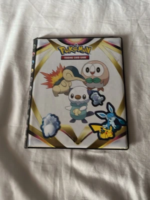 Pokémon samlarkortspärm - Säljer en Pokémon samlarkortspärm med färgglad framsida som har motiv av flera populära Pokémon, bland annat Pikachu, Rowlet och Cyndaquil. Pärmen är vit med guldiga detaljer och har plats för många kort. Perfekt för att organisera och visa upp din samling.