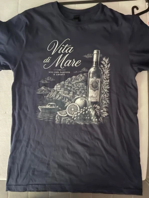 Vita di Mare t-shirt med tryck - Snygg t-shirt med 'Vita di Mare'-tryck framtill. Motivet visar en vinflaska, frukt och kustlandskap i vitt på marinblå eller vit botten. Klassisk rund hals och korta ärmar. Perfekt för dig som gillar stilrena och grafiska prints.