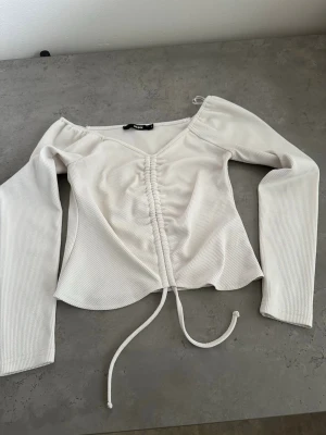 Vit ribbad topp med snörning - Superfin vit ribbad topp med lång ärm och v-ringning. Toppen har snörning framtill som ger en snygg detalj och croppad passform. Perfekt att matcha med jeans eller kjol för en trendig look.