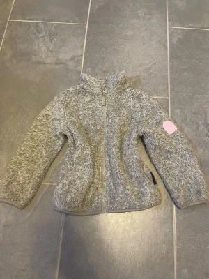 Grå teddyjacka med rosa detalj - Mysig grå teddyjacka med dragkedja framtill och hög krage. Jackan har en rosa patch på ärmen och är tillverkad i fluffigt, mjukt material som håller dig varm. Perfekt för kyliga dagar och enkel att matcha med olika outfits.