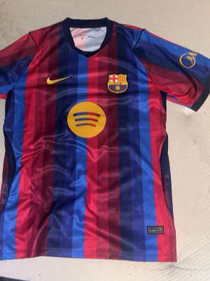 Barcelona matchtröja randig Nike - Snygg Barcelona matchtröja med vertikala blå och röda ränder, officiella klubbmärket och Nike-logga på bröstet samt Spotify-logga i gult. Tillverkad i lätt och ventilerande material, perfekt för fotboll eller träning. BARCA FOUNDATION-tryck på ryggen.