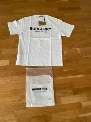 Vit Burberry t-shirt  - Säljer en vit t-shirt från Burberry med stor svart logga och texten 'London England' på bröstet. Klassisk rund halsringning och korta ärmar. Tillverkad i mjuk bomull och levereras med originalförpackning och tagg. Denna t shirt och den svarta jag även har kan båda köpas för 699 som ett paket. Tröjorna är i storlek S men passar mera M.