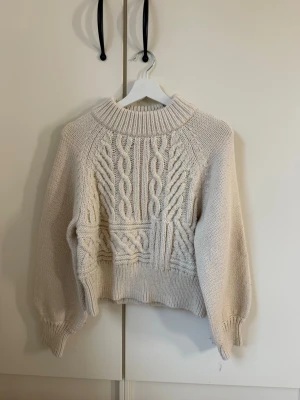 Beige kabelstickad tröja med polokrage - Säljer en beige kabelstickad tröja med polokrage och breda muddar. Tröjan har ett snyggt mönster framtill med flätstickning och är gjord i ett mjukt material. Perfekt för kyliga dagar och passar till det mesta i garderoben.