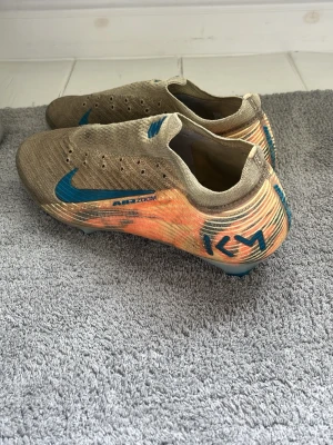 Nike fotbollsskor Air Zoom orange/blå - Snygga fotbollsskor från Nike med Air Zoom-teknologi. Skorna är beige med orange och blå detaljer, inklusive en blå Nike-swoosh och KM-logga på hälen. Ovandelen är i stickat material för bra passform och grepp. Sulan har blå dobbar och genomskinliga partier.