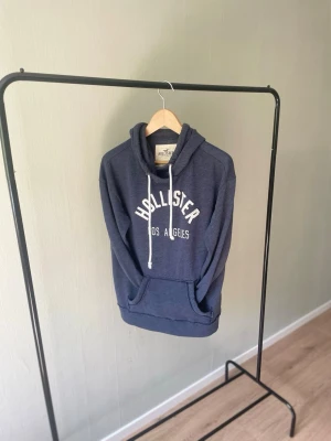 Hollister Hoodie - Hollister hoodie – Strl M 🔥 Mörkblå hoodie från Hollister med stort logotryck framtill. Mjuk och bekväm modell med huva, dragsnören och känguruficka – perfekt för en avslappnad vardagslook. I fint begagnat skick. 📏 Storlek: M 📦 Skick: Bra begagnat skick 💬 Hör gärna av dig vid frågor eller om du vill ha fler bilder!