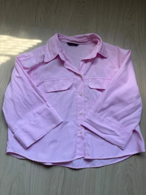 Ljusrosa skjorta från Zara - Säljer en ljusrosa skjorta från Zara med klassisk krage, knäppning framtill och två bröstfickor med lock. Skjortan har lång ärm och är tillverkad i ett mjukt material som känns skönt mot huden. Perfekt för en avslappnad och trendig look.