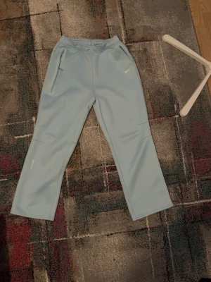 Ljusblå mjukisbyxor från NOCTA - Snygga ljusblå mjukisbyxor från NOCTA med raka ben och diskret Nike-logga på ena höften. Byxorna har dragkedjefickor och elastisk midja för skön passform. Perfekta för en chill och trendig look.