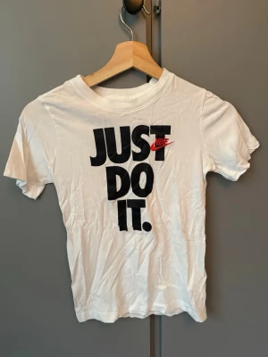 Vit Nike Just Do It t-shirt - Vit t-shirt från Nike med det klassiska 'Just Do It.' trycket i svart och rött framtill. T-shirten har rund halsringning och korta ärmar. Tillverkad i mjuk bomull för en skön och avslappnad stil.
