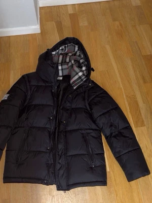 Svart pufferjacka med huva - Säljer en stilren svart Burberry-jacka i exklusiv design. Perfekt för dig som gillar en clean och klassisk look med premiumkänsla. Passar både till vardags och mer uppklädda outfits. Storlek L men passar lika med M