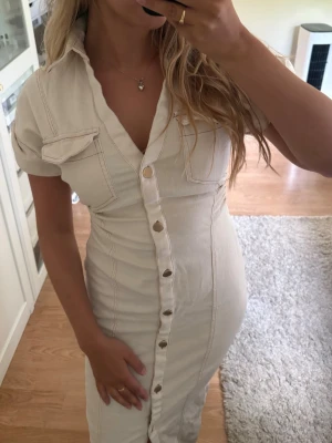 Beige långklänning - Snygg beige jeans långklänning med korta ärmar och markerad midja/figursydd. Klänningen har v-ringning, framfickor på bröstet och stora dekorativa knappar längs hela framsidan. Denna klänning är slutsåld på hemsidan, använd 1 gång!! Nypris 499kr❤️